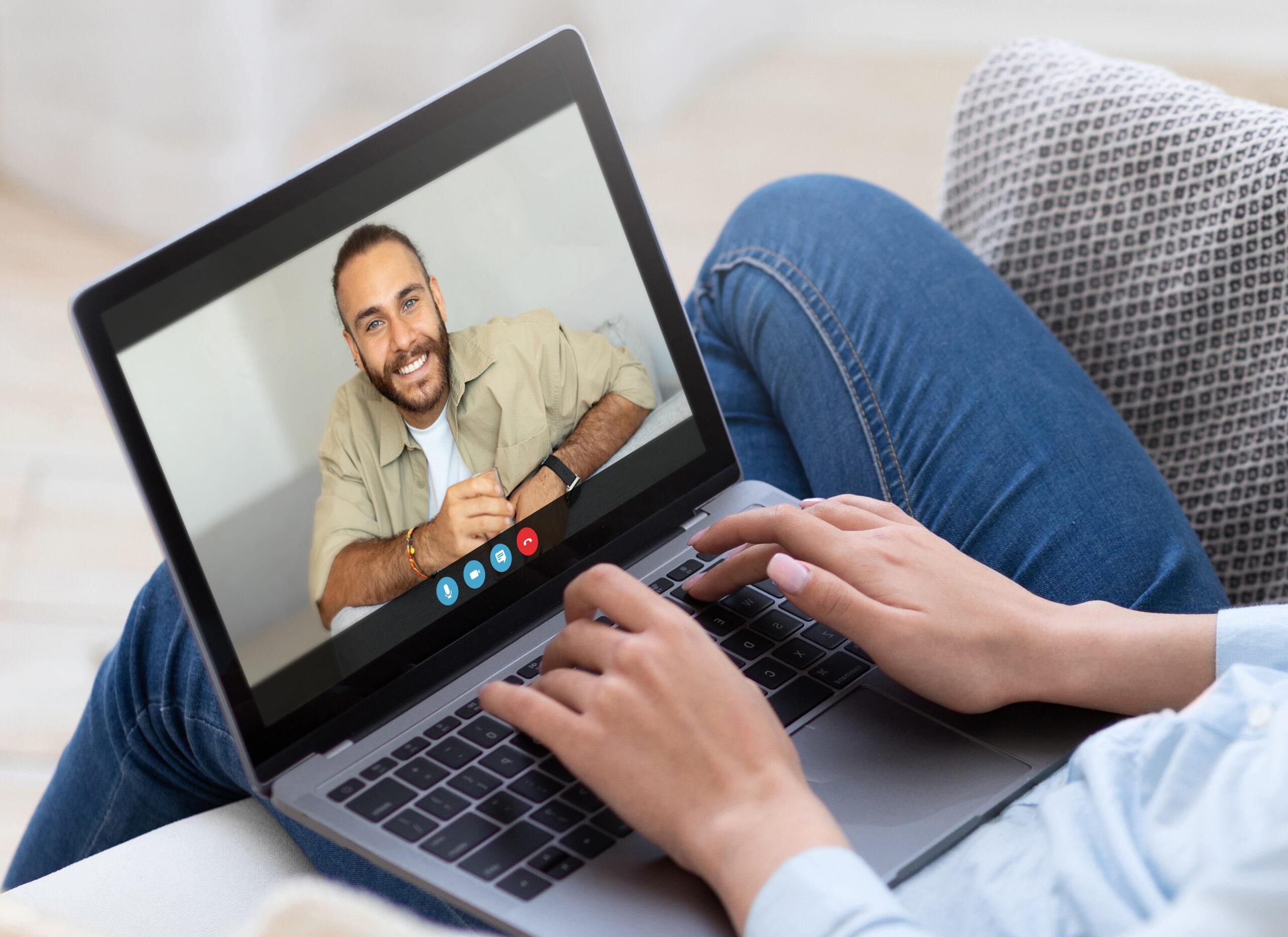 A woman is sitting on a couch and video chatting with a man on a laptop computer. She is typing on the keyboard and looking at the screen. The man is smiling and appears to be talking.